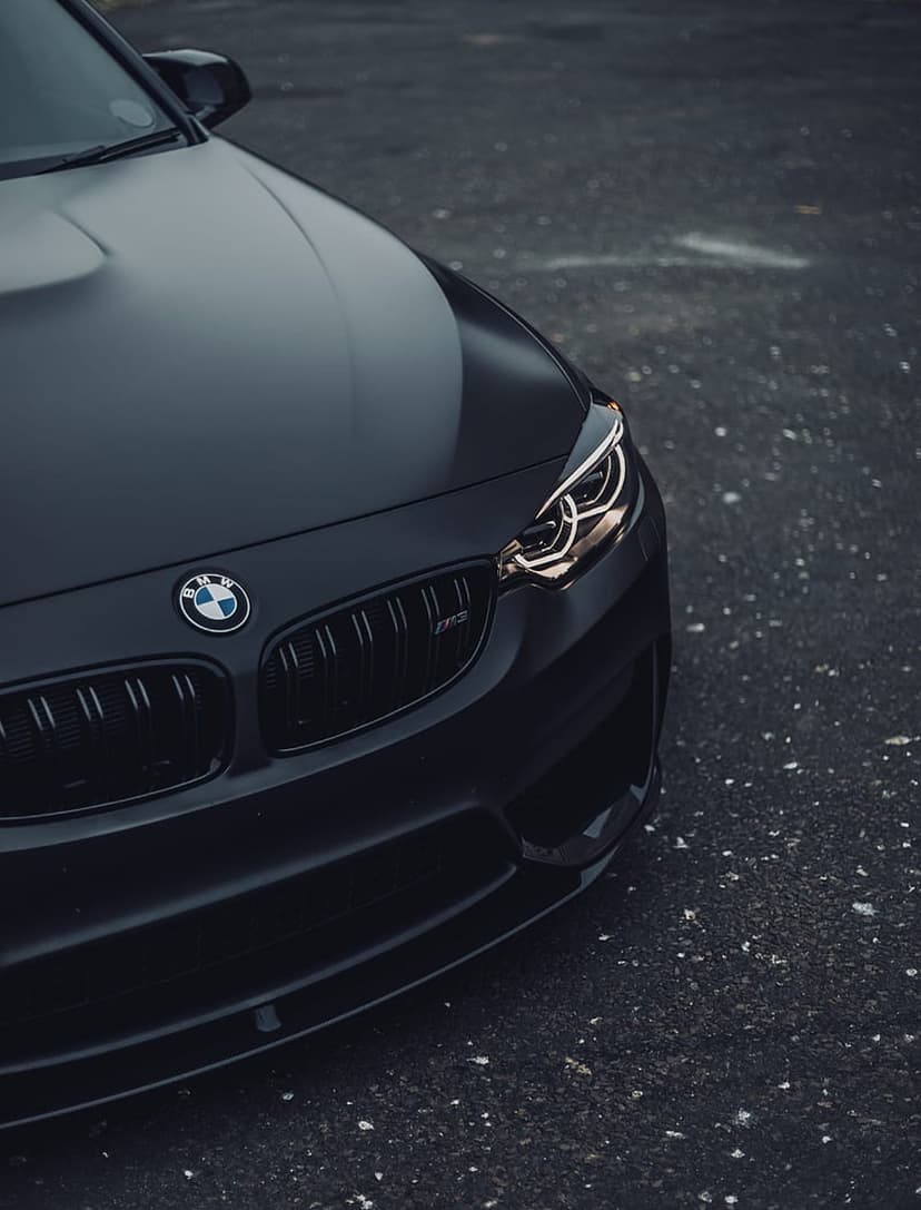Black BMW M car with glowing LED headlights, matte hood, and signature kidney grille on a dark asphalt surface.
