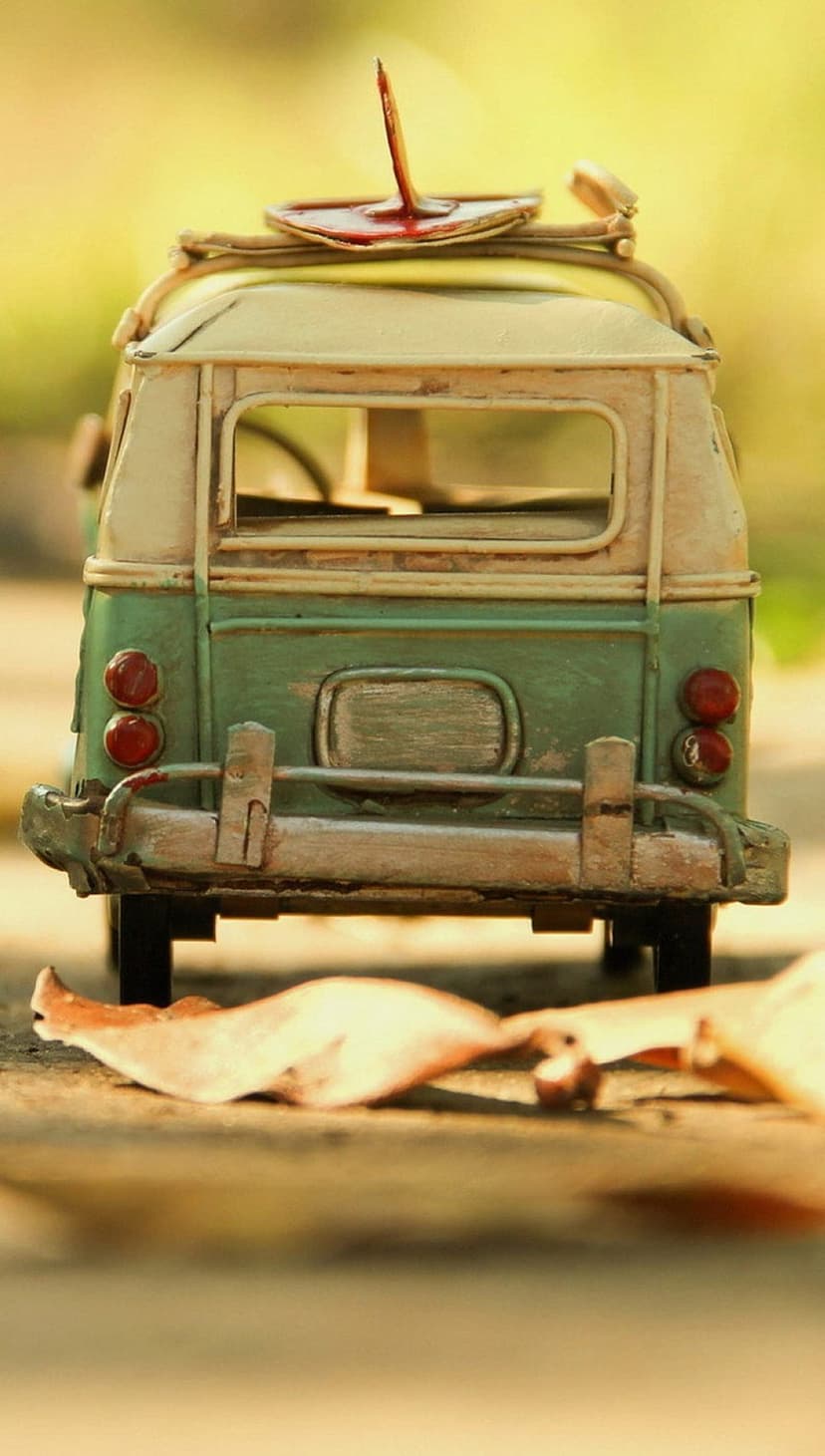 Green vintage toy van with a rusty nail in the roof sits among orange autumn leaves in a blurred forest.