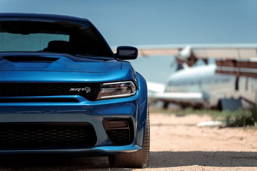Blue Dodge Charger SRT Hellcat parked on a sandy airfield with a blurred propeller airplane background.