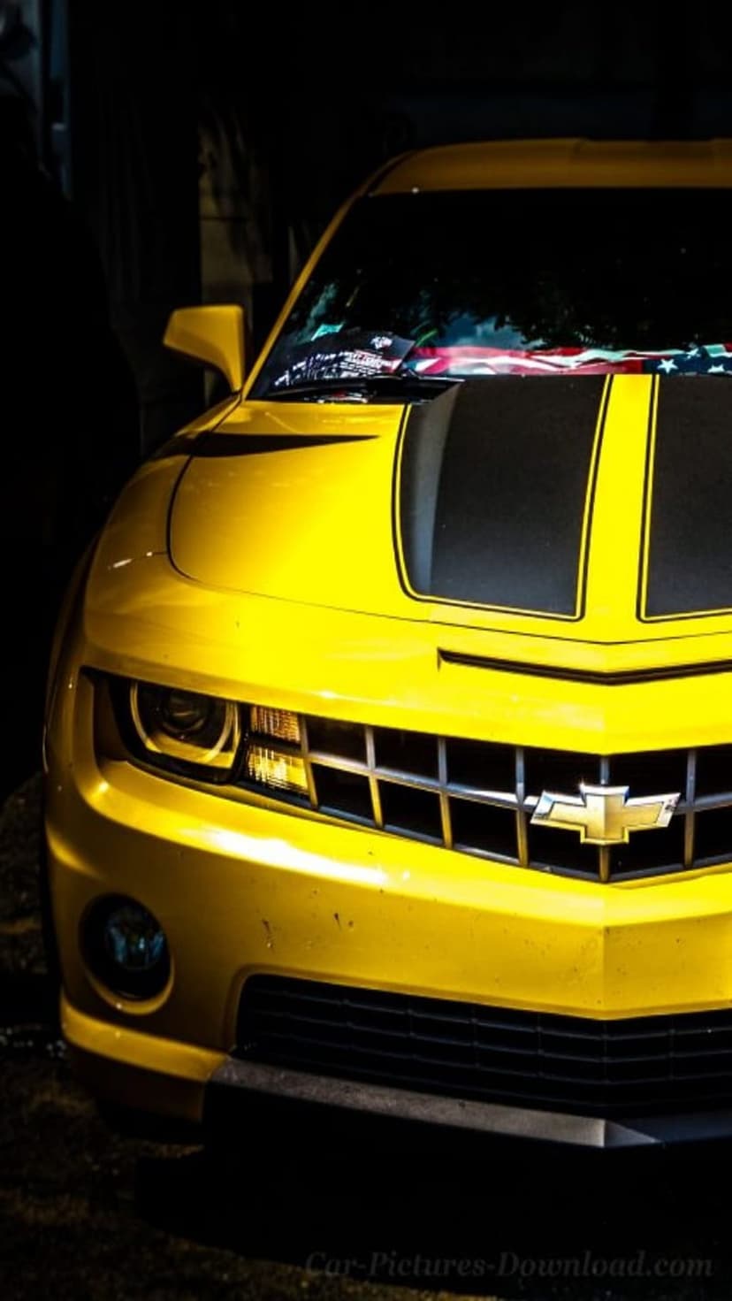 Yellow Chevrolet Camaro front grille with black racing stripes and lit headlights against a dark background.