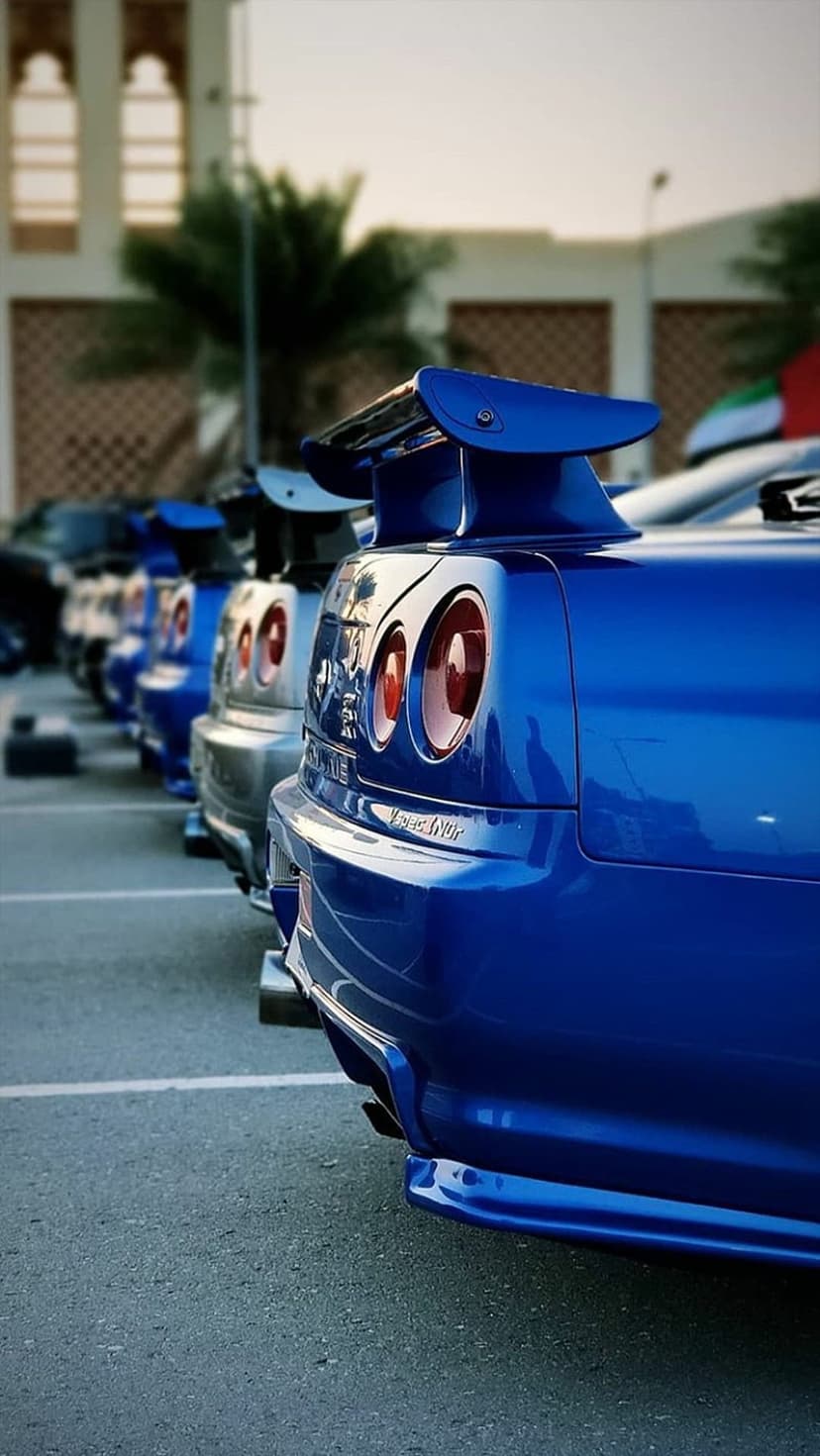 Blue Nissan Skyline R34 rear wing and circular taillights parked outdoors near palm trees in bright daylight.