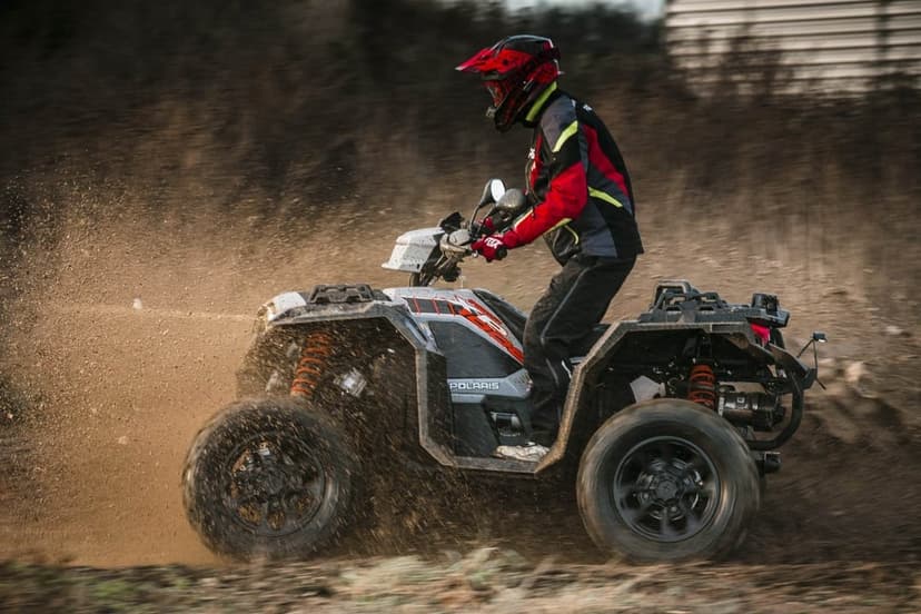 A Polaris ATV kicks up dirt in a powerful drift with a rider in red and black gear against a blurred background.