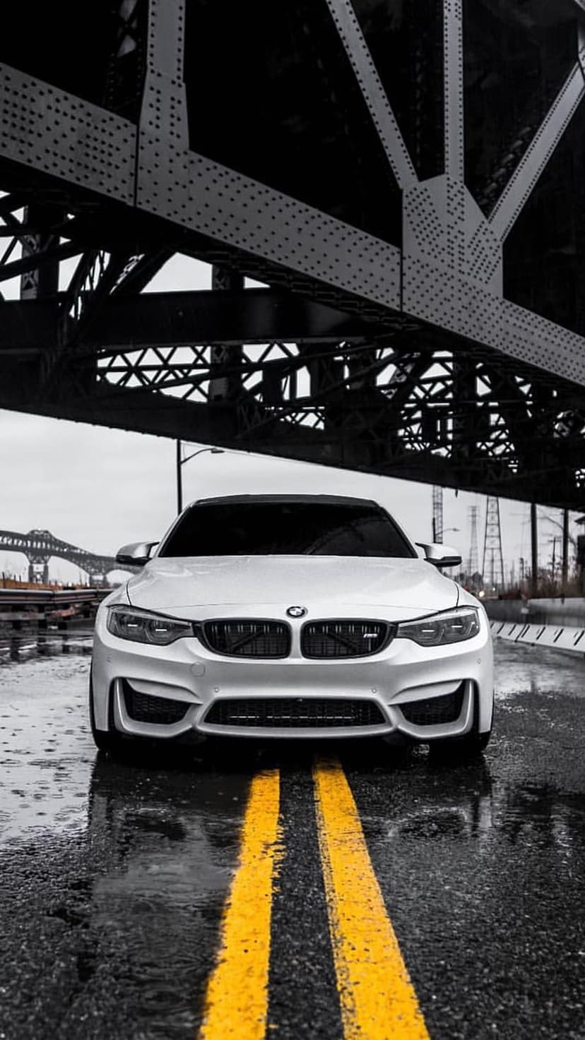 White BMW M4 parked beneath a heavy steel bridge on a wet road with yellow lines in an urban industrial area.