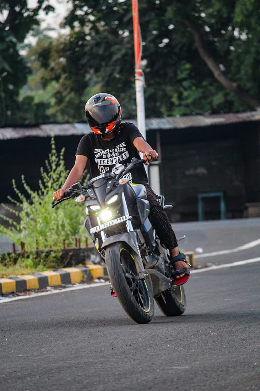 Motorcyclist in a helmet and black t-shirt performs a high wheelie on a street with trees and a building behind.