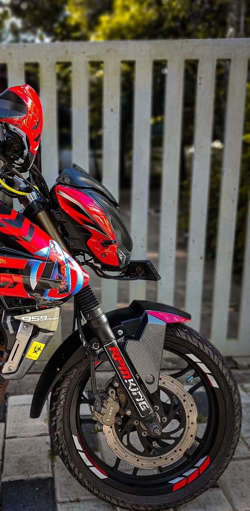 Custom red and black Avengers motorcycle with chrome forks stands before a blurred green garden background.