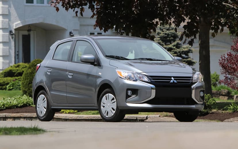 Grey Mitsubishi Mirage hatchback parked on a paved driveway in front of a white house with a leafy tree.