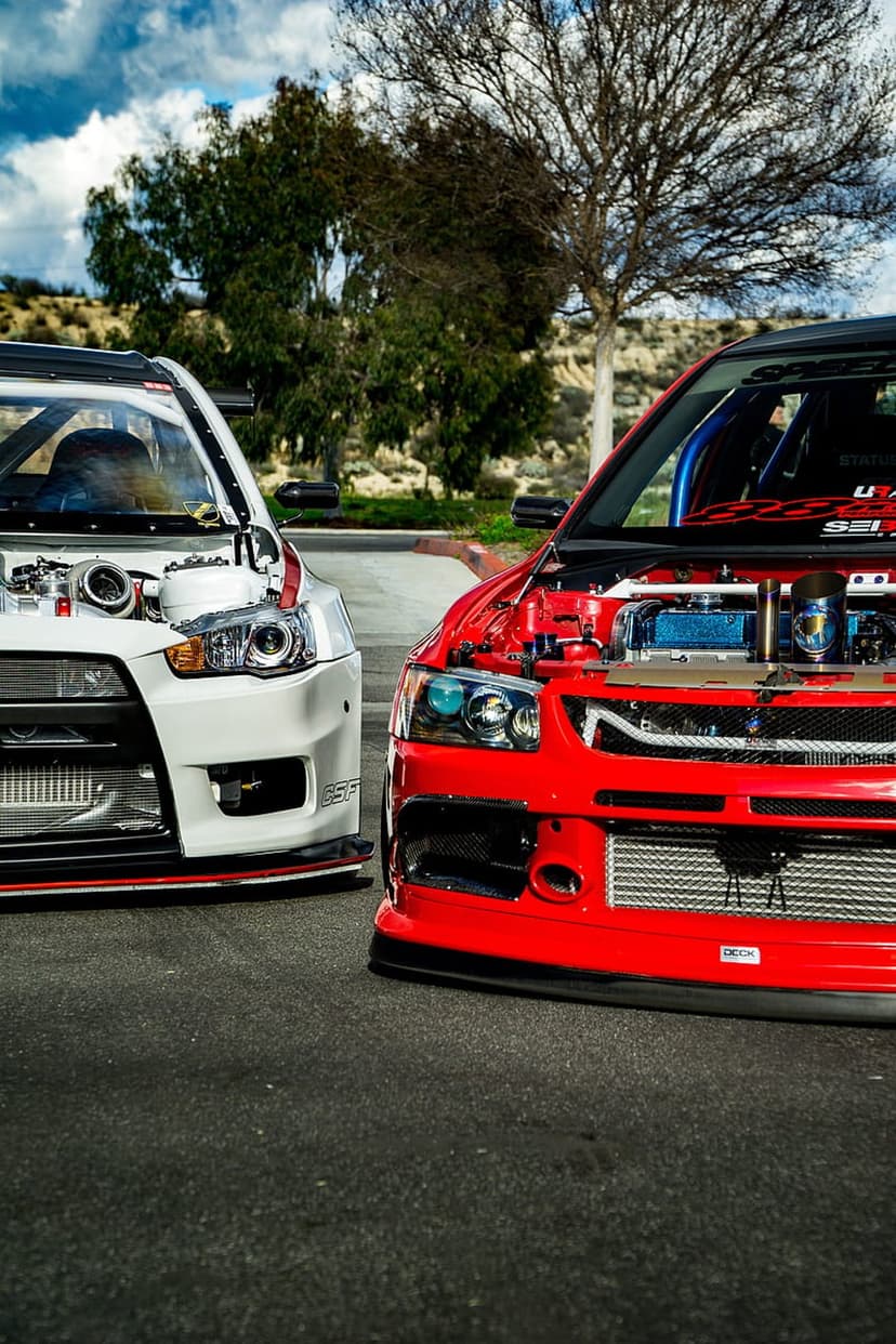 Red and white Mitsubishi Lancer Evolution cars with exposed engines parked outdoors under a cloudy sky.