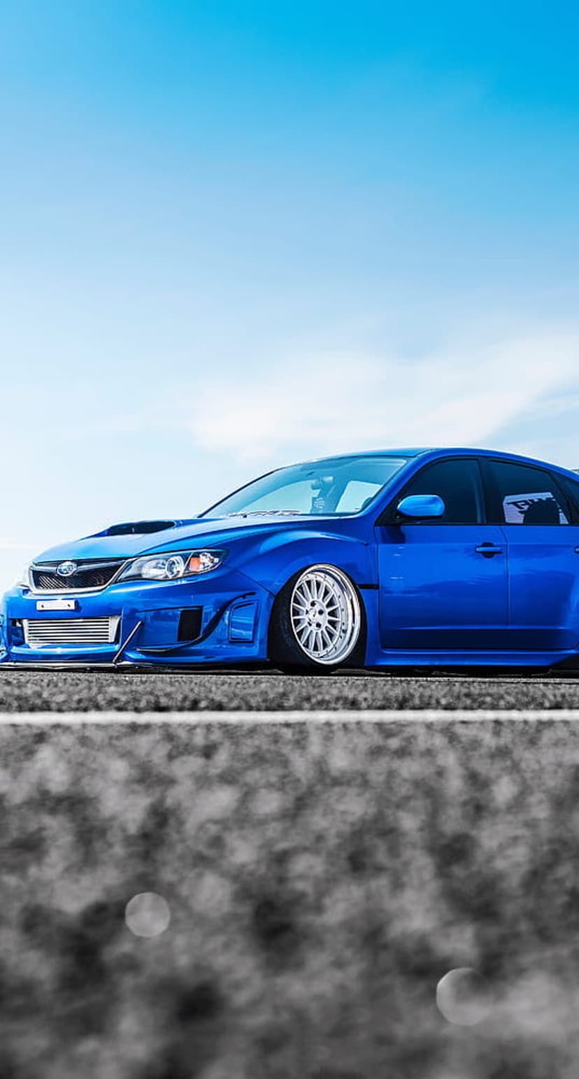 Blue Subaru Impreza STI with widebody kit and silver wheels parked on asphalt under a bright cloudy sky.
