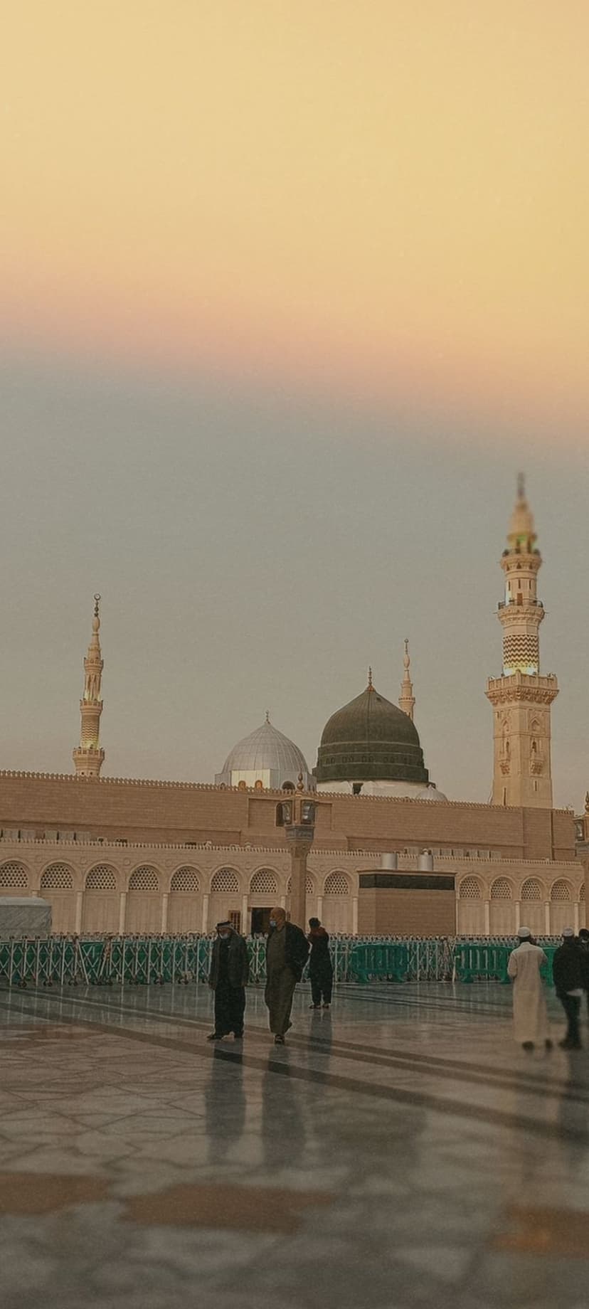 People walk across a polished mosque courtyard toward minarets and domes under a vibrant sunset sky.