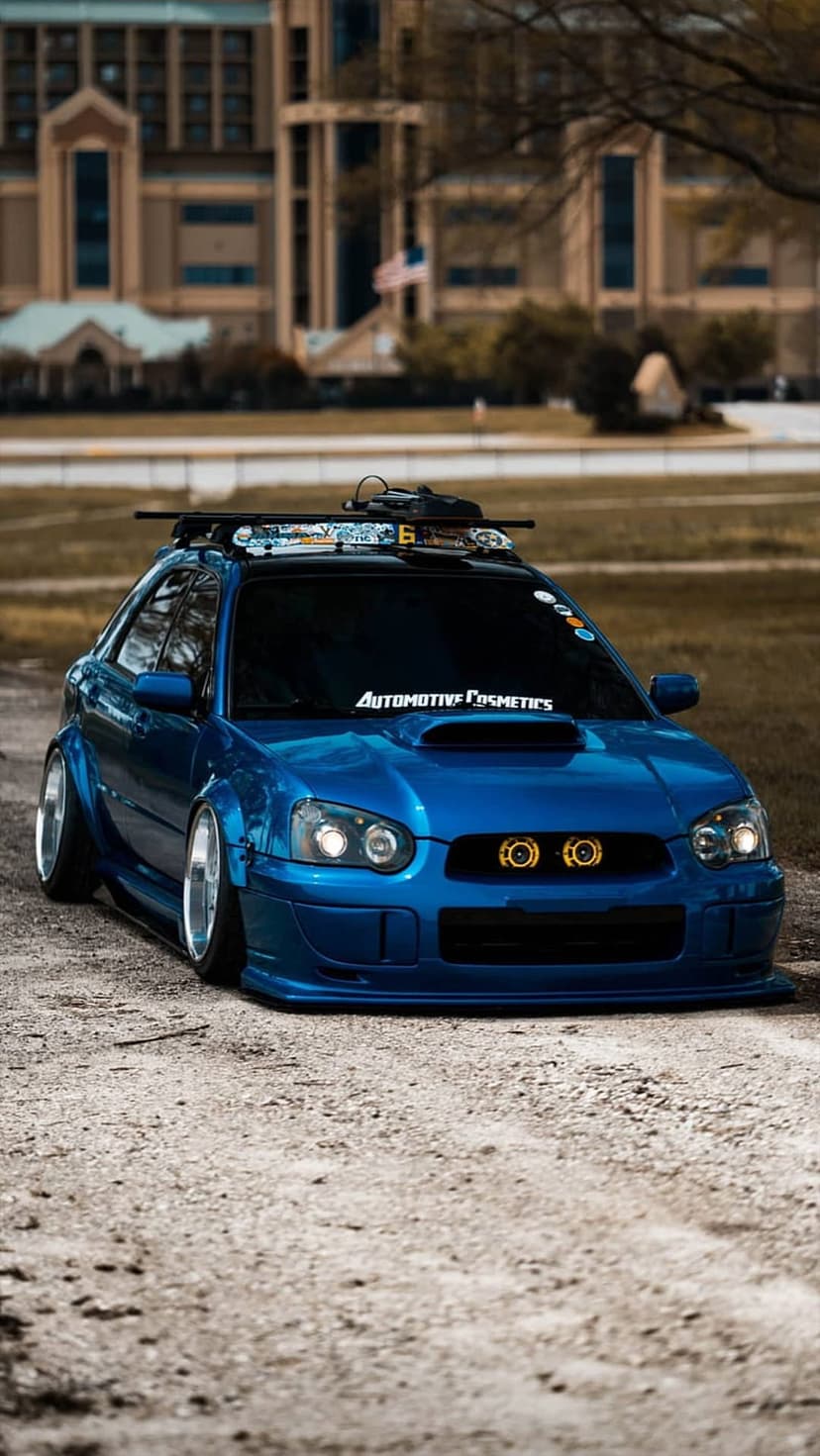 Blue Subaru WRX STi with a widebody kit and roof rack parked on a dirt road with a blurred background.
