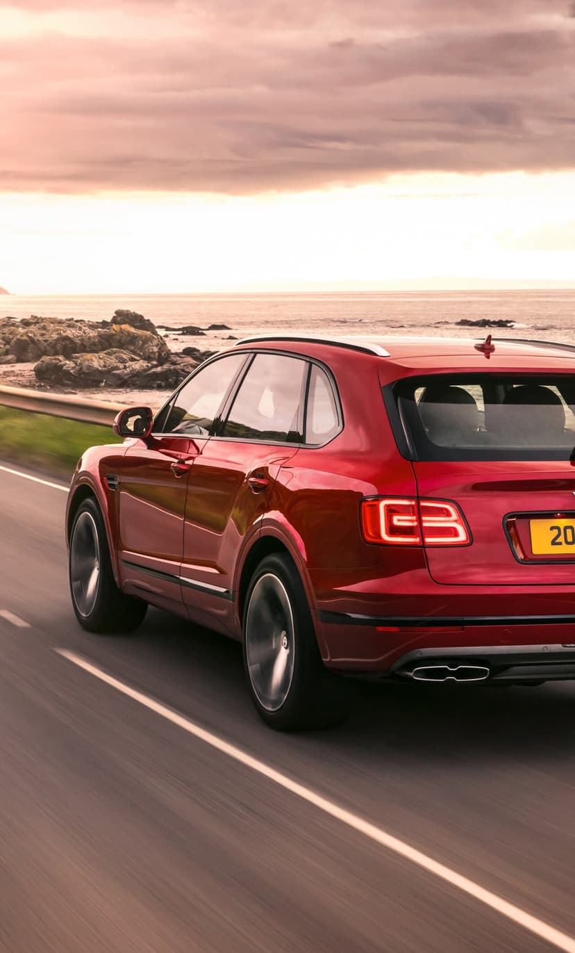 Red luxury SUV driving on a coastal road at sunset with glowing taillights and ocean views.
