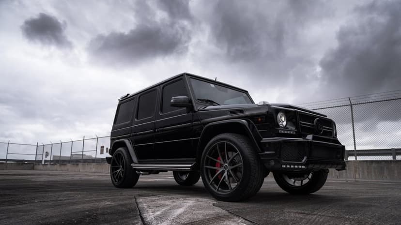 Black Mercedes-Benz G-Class Brabus SUV with red brake calipers parked under a dramatic cloudy sky on concrete.