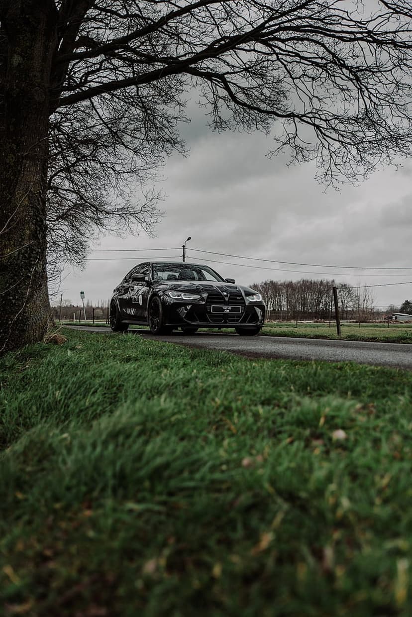 Black BMW M3 sedan parked on an asphalt road under a dark, overcast sky with green grass and bare tree branches.