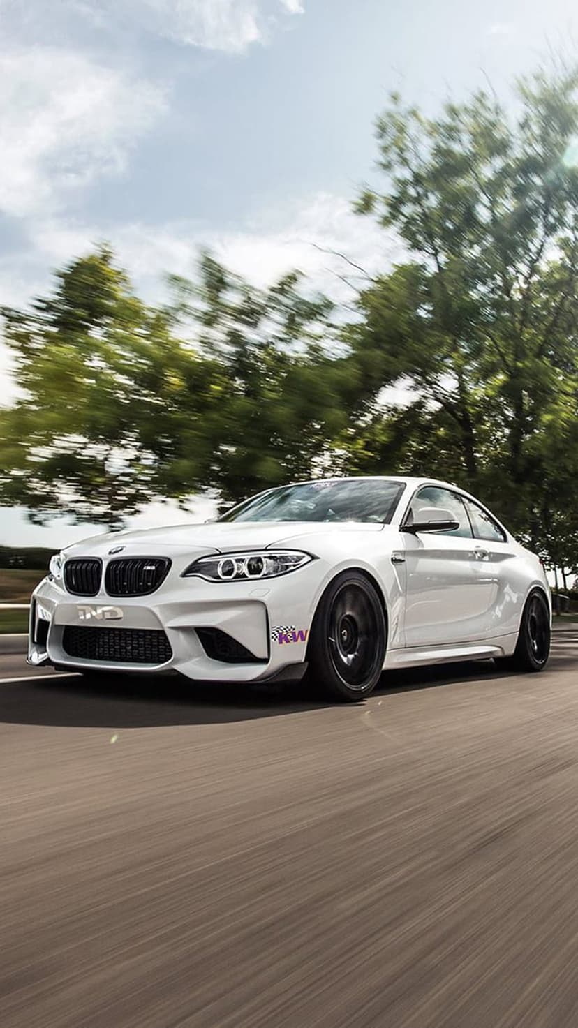White BMW M2 coupe driving on a paved road with black wheels, motion blur, and a background of trees and sky.