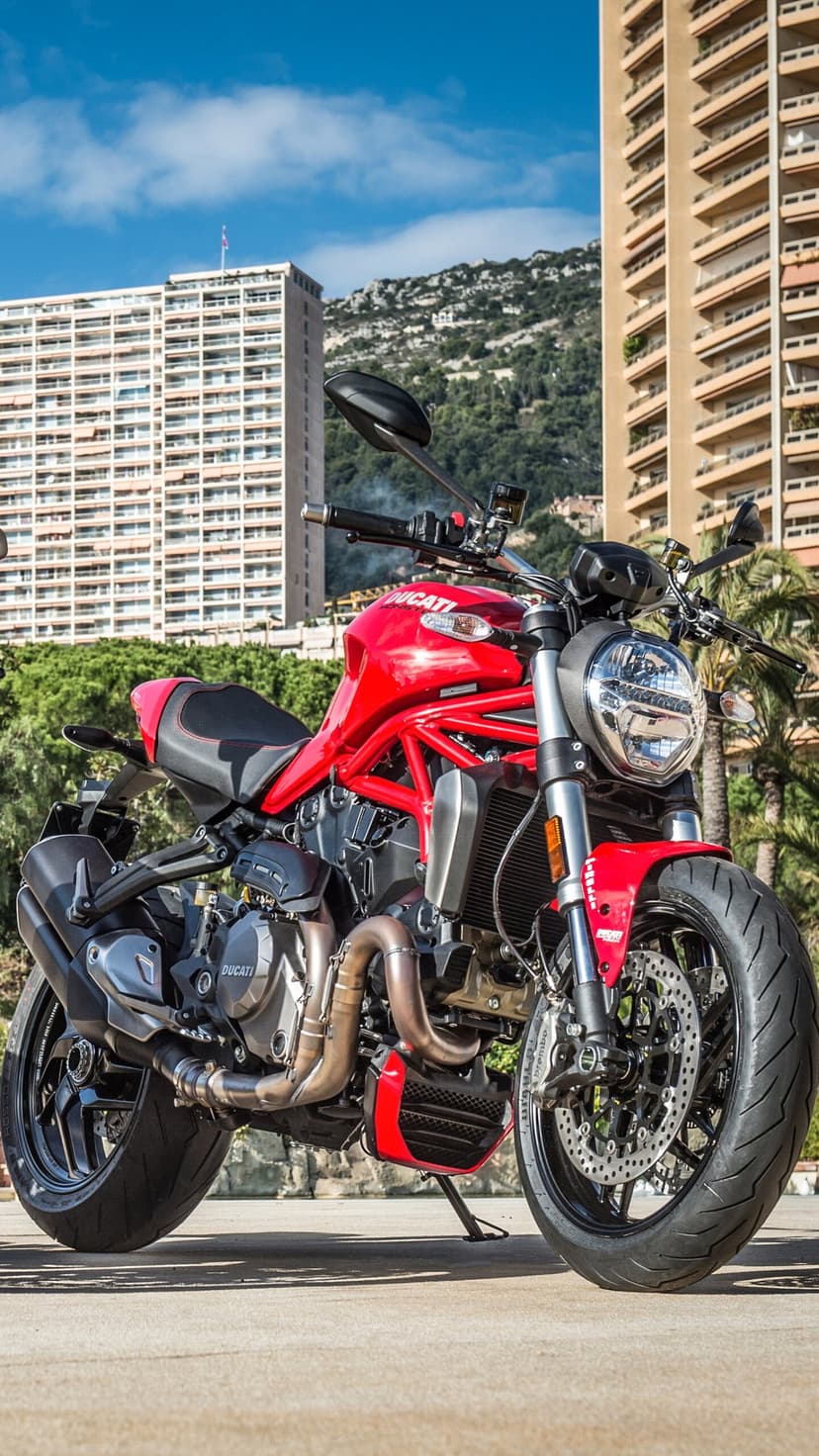 Red Ducati Monster motorcycle with exposed engine parked in front of tall city buildings and green hills.