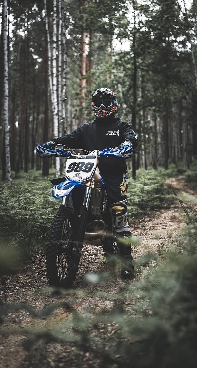 Motocross rider in full gear stands beside a dirt bike on a fern-covered trail under towering forest trees.