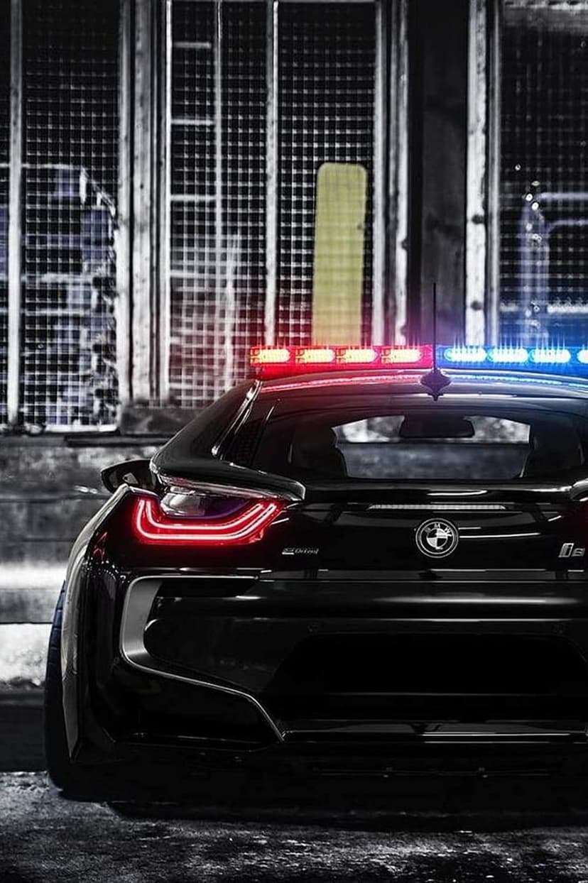 Black BMW police car with flashing red and blue lights parked against a grey industrial wall at night.