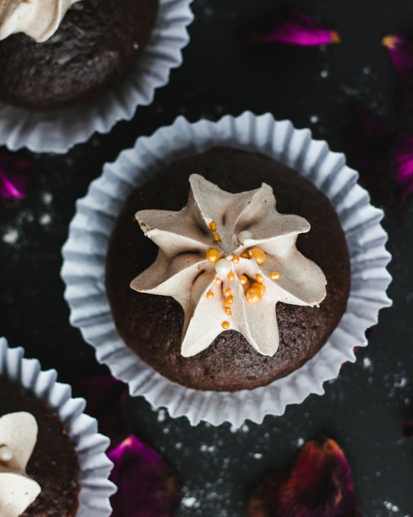 Chocolate cupcakes with swirled mocha frosting and gold sprinkles sit on a dark surface with purple petals.