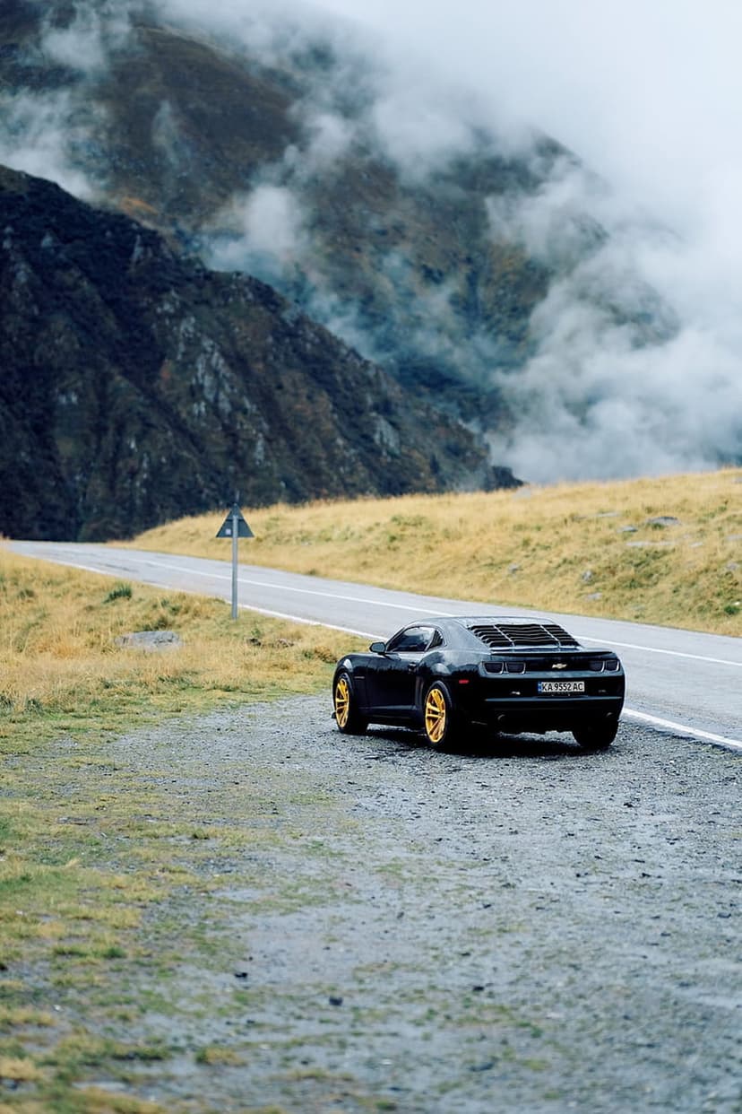 Black Chevrolet Camaro with gold rims parked on a winding road among foggy mountains and golden grass fields.