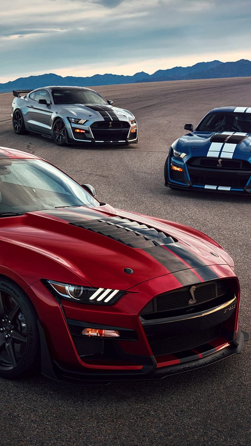 Red, blue, and silver Ford Mustang Shelby GT500 cars with racing stripes parked on asphalt near mountains.