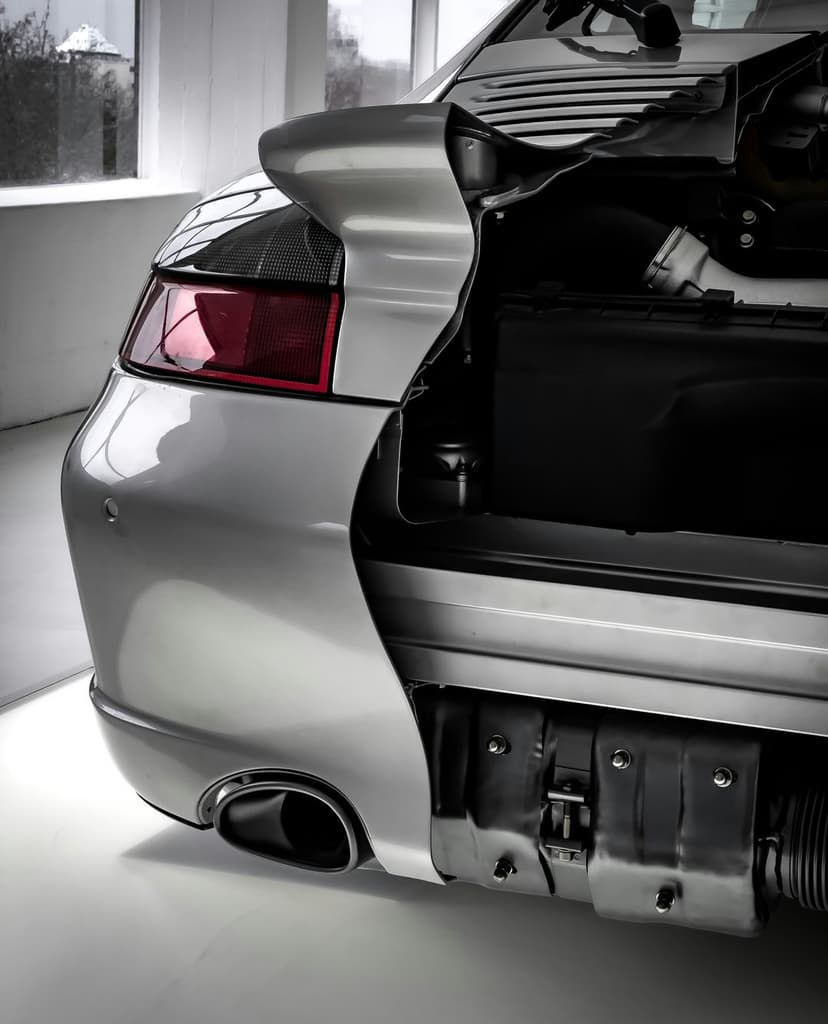 Silver Porsche rear with an open engine bay showing metallic parts and a red taillight on a white surface.