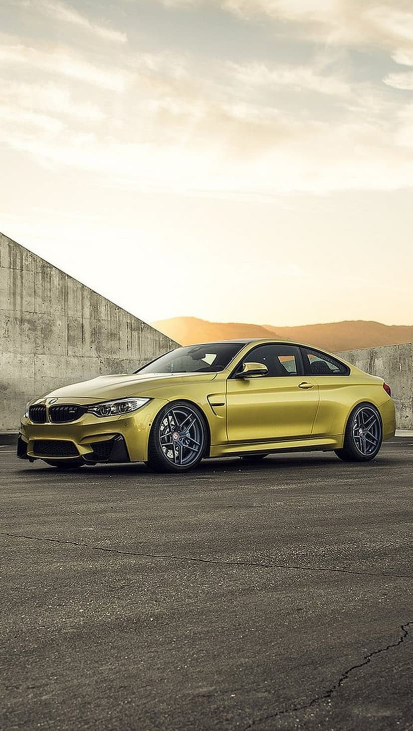 Yellow BMW M4 Coupe with dark rims parked on cracked asphalt against a concrete wall under a sunset sky.