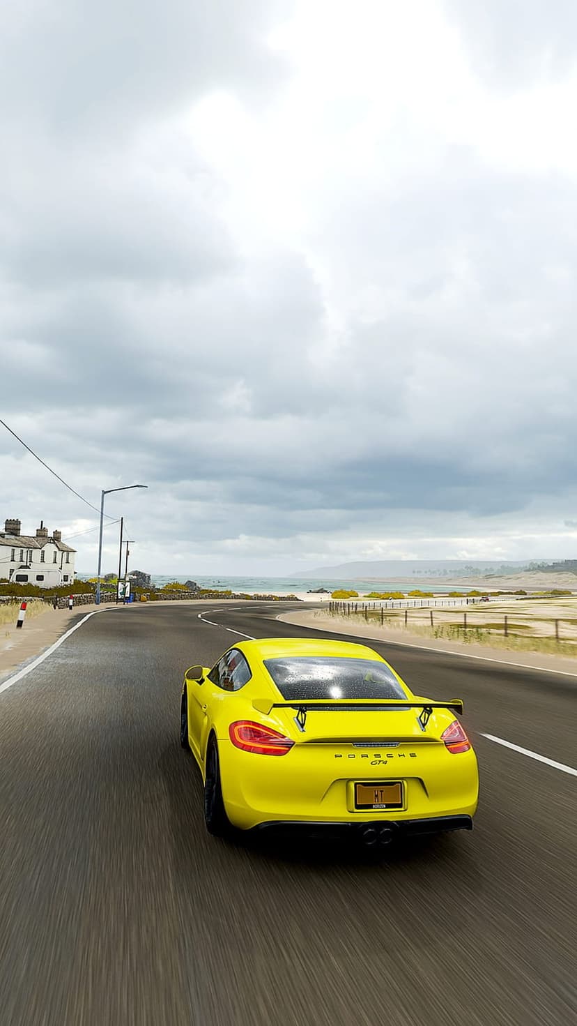 Yellow Porsche Cayman GT4 drives on a damp seaside road under a moody gray sky beside a sandy beach.