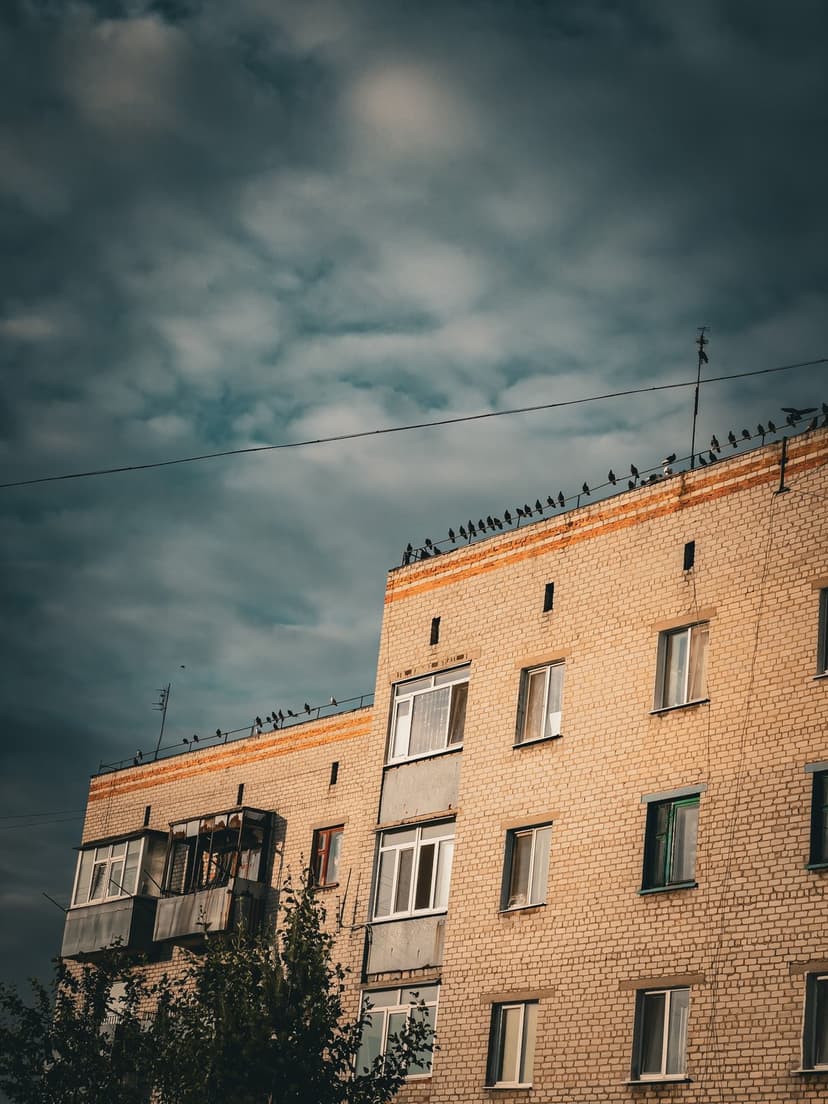 Pigeons perch along a light brown brick apartment ledge under a vast expanse of heavy, grey storm clouds.