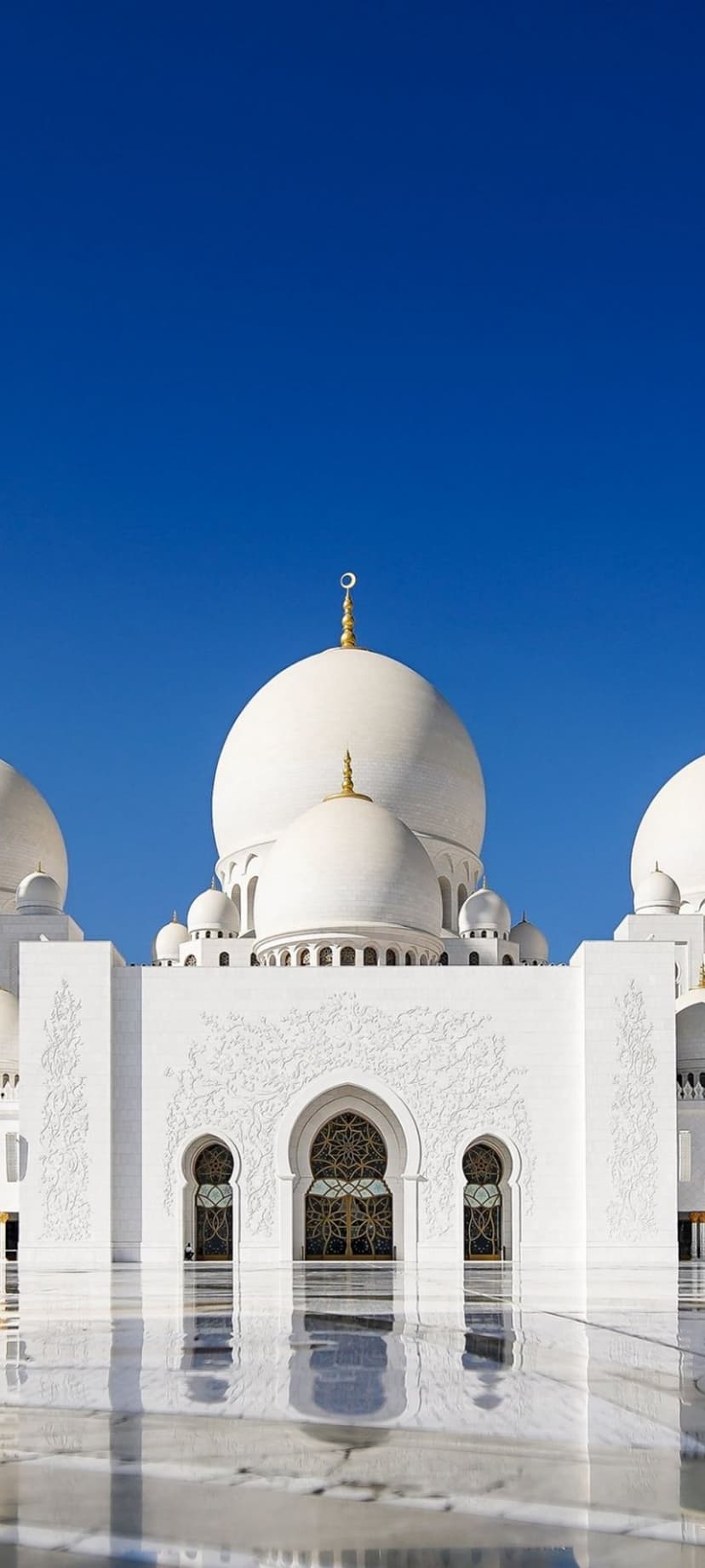 Sheikh Zayed Grand Mosque white domes reflect on a wet marble floor under a clear blue sky in Abu Dhabi.