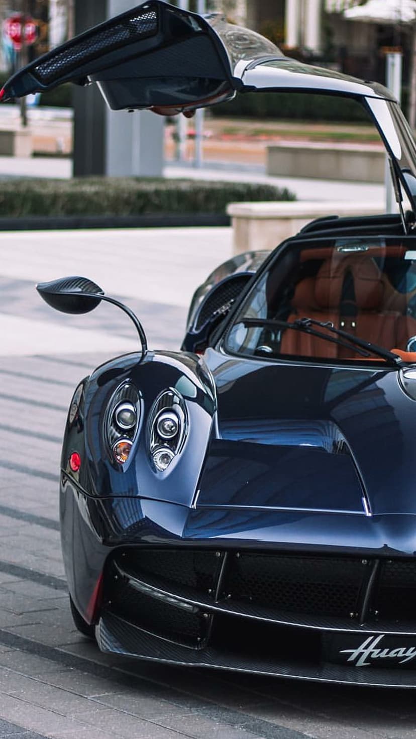 Dark blue Pagani Huayra with open butterfly doors, tan leather interior, and carbon fiber headlight detailing.
