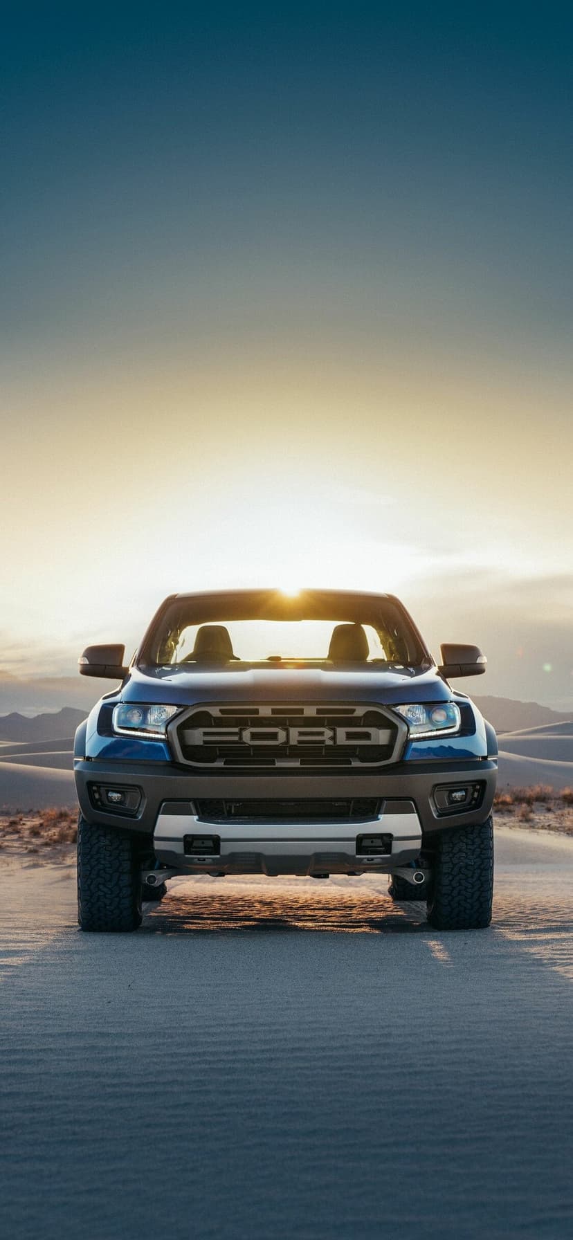 Blue off-road pickup truck centered on sand dunes with sun flare and a vast desert background at golden hour.