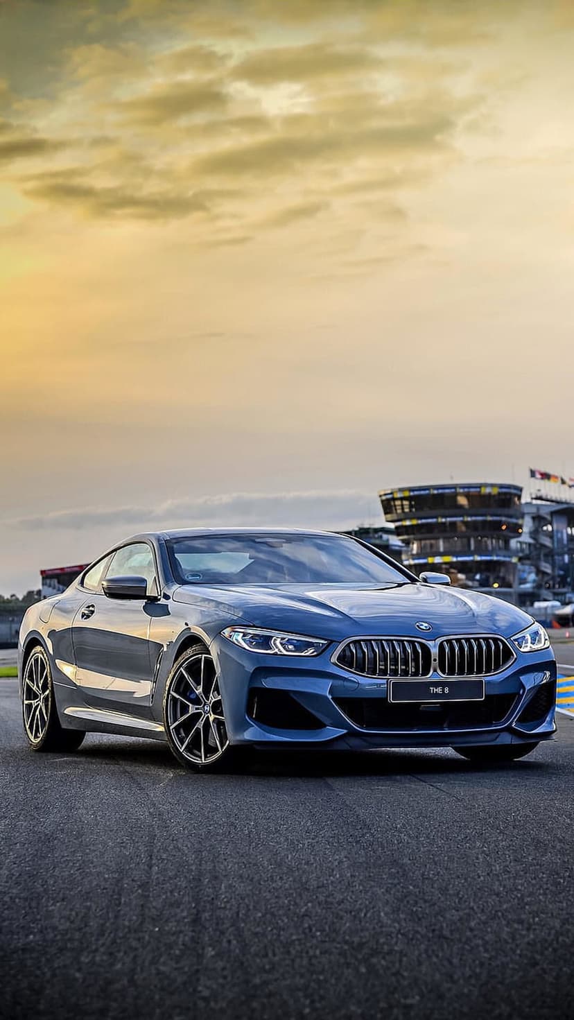 Blue BMW M8 Coupe on an asphalt racetrack at sunset with a hazy sky and modern racing facility in the background.