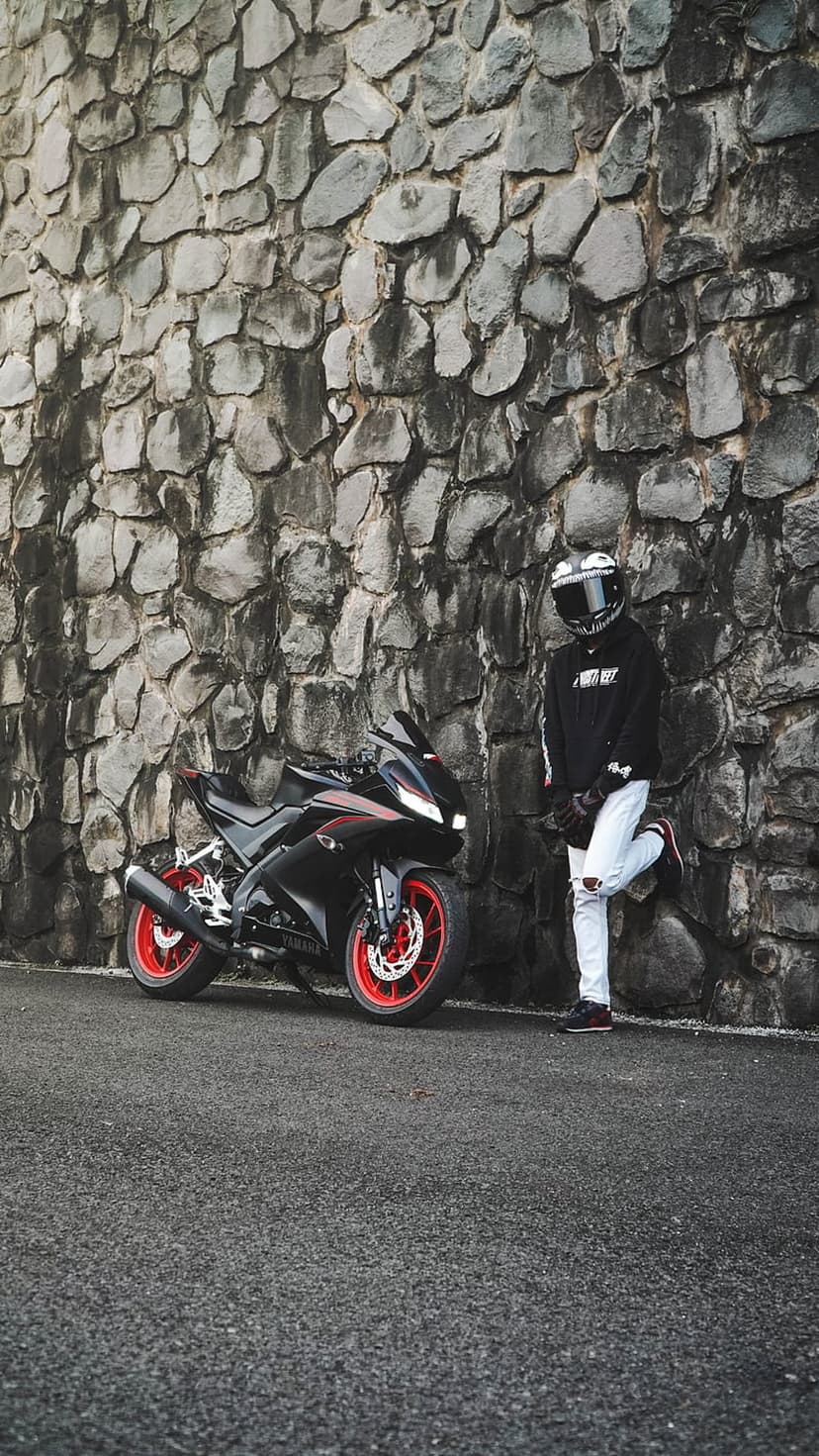 Biker in a black hoodie and white ripped jeans leans against a stone wall beside a black and red sportbike.