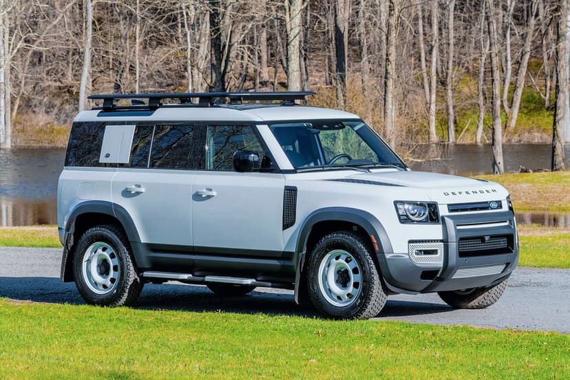 White Land Rover Defender 110 SUV with a roof rack parked on a gravel path near a green lawn and scenic lake.