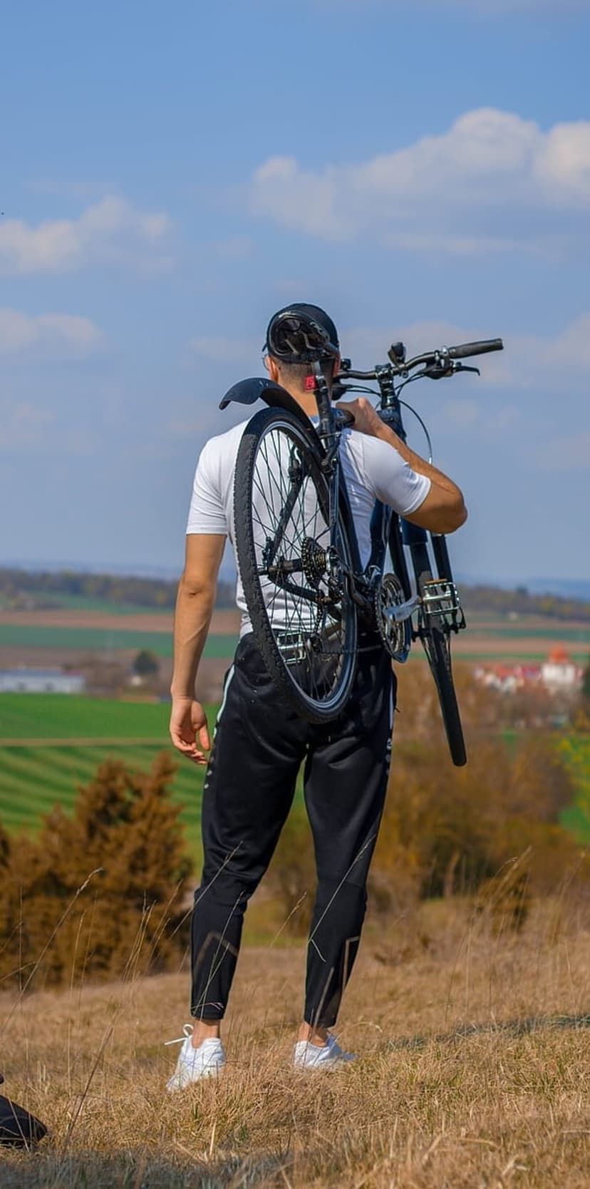Man in athletic clothing carries a bicycle over his shoulder while walking through a vast hilly green landscape.