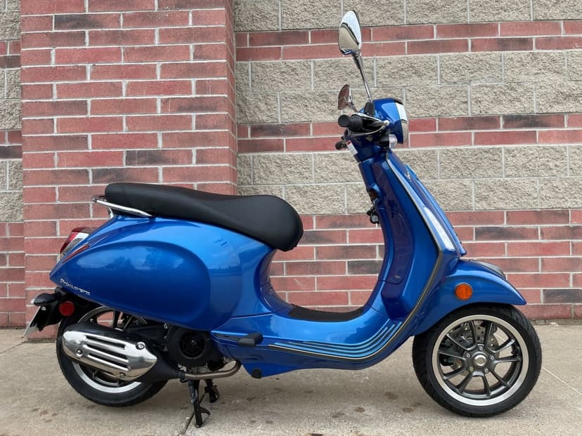Blue Vespa Primavera scooter parked against a brick wall, showing chrome mirrors and metallic paint finish.