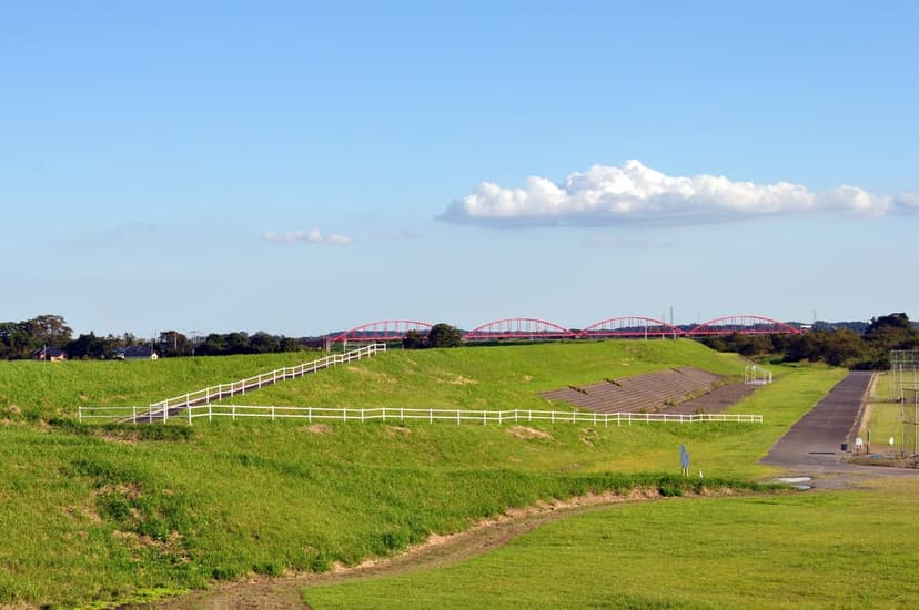 Red arched bridges and a white fence sit atop a vibrant green hillside under a clear blue sky with soft clouds.