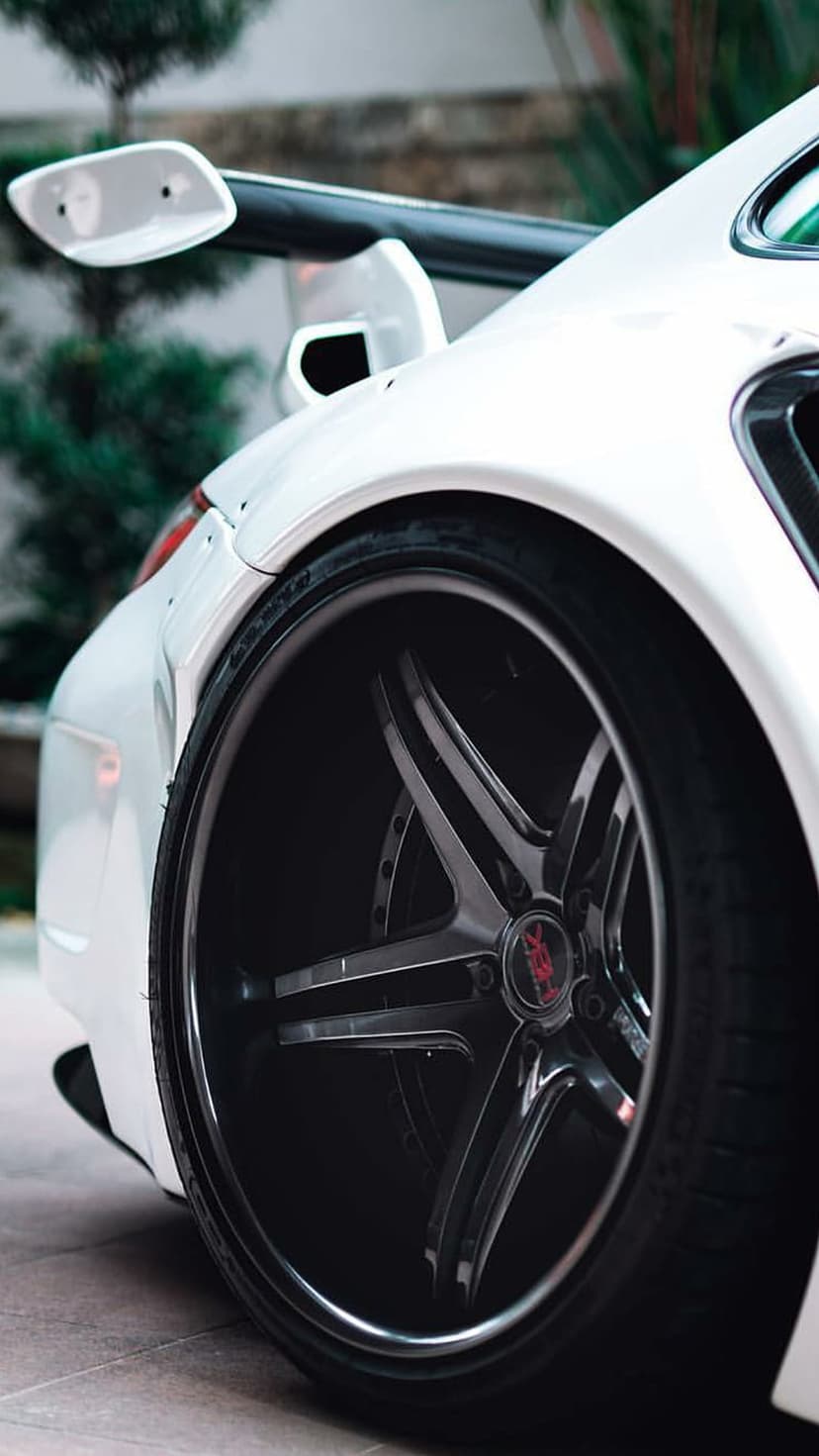 White Porsche sports car featuring a black five-spoke wheel and aerodynamic rear spoiler against green plants.