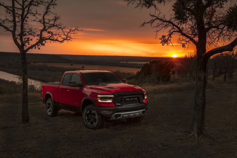 Red Ram 1500 Rebel pickup truck on a grassy ridge framed by silhouetted trees during a vibrant orange sunset.