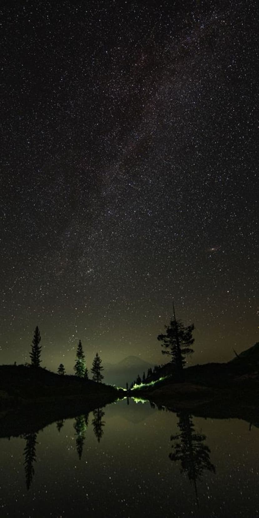 Milky Way galaxy core reflected in a still lake at night with evergreen tree silhouettes on the shore.