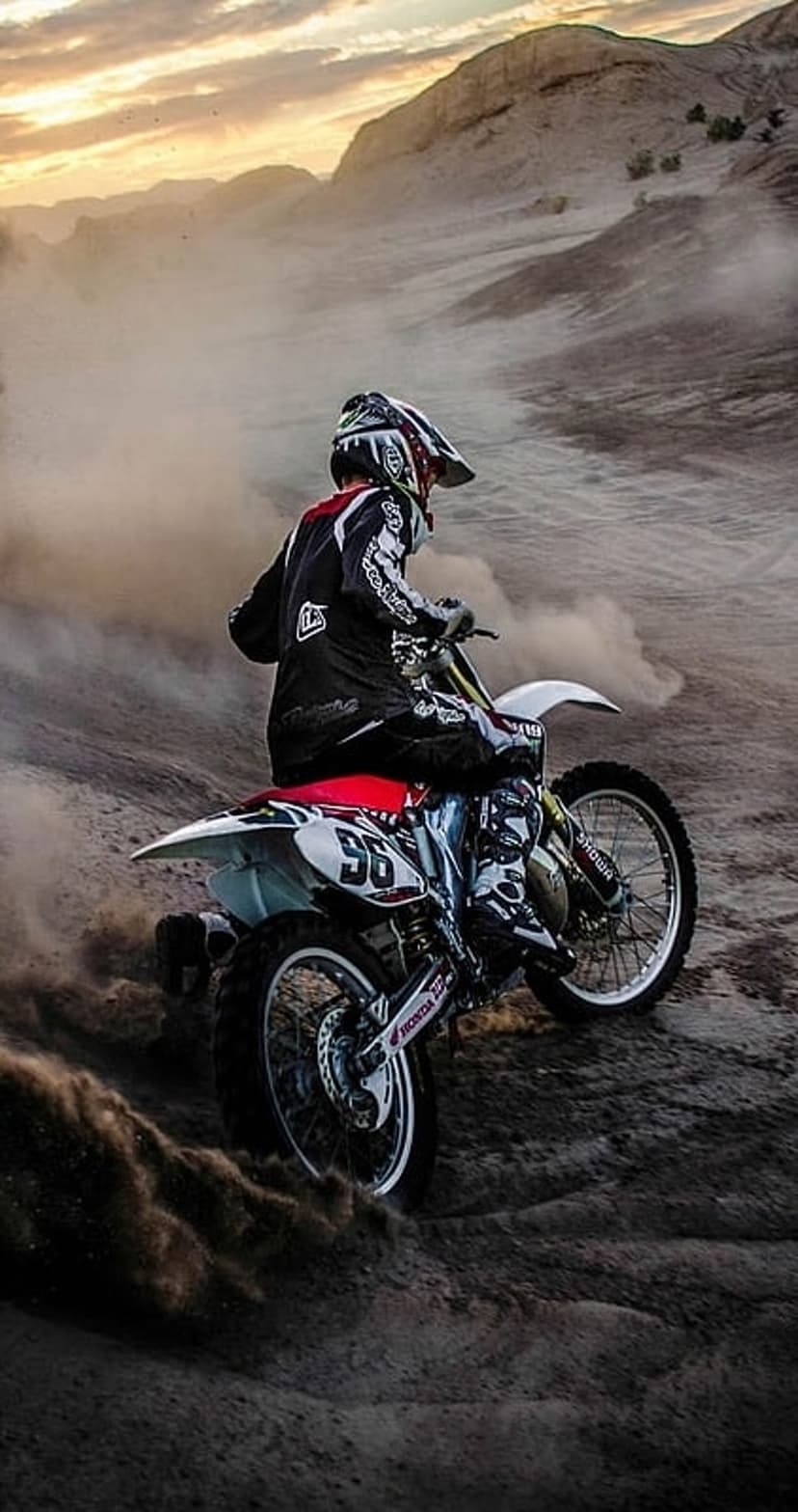 Motocross rider in full protective gear maneuvers a dirt bike through clouds of dust on a sandy desert ridge.