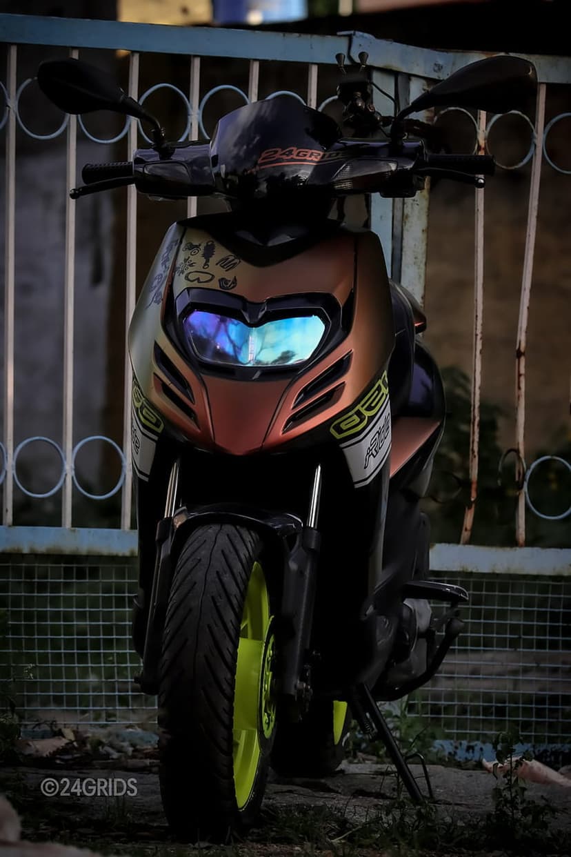 Custom bronze scooter with neon green wheels and a glowing blue headlight parked in front of a metal fence.
