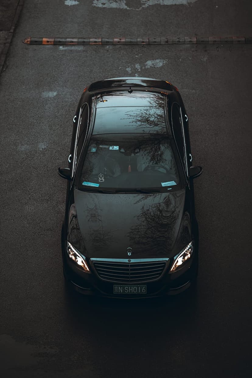 Black Mercedes S-Class luxury sedan parked on wet asphalt seen from a high overhead bird's-eye perspective.