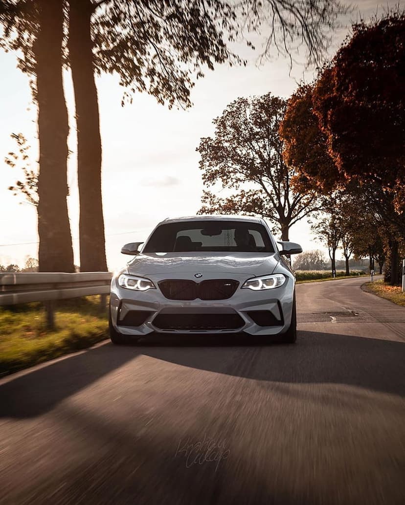 BMW M2 silver sports car speeding on a road lined with orange autumn trees during a bright golden hour sunset.