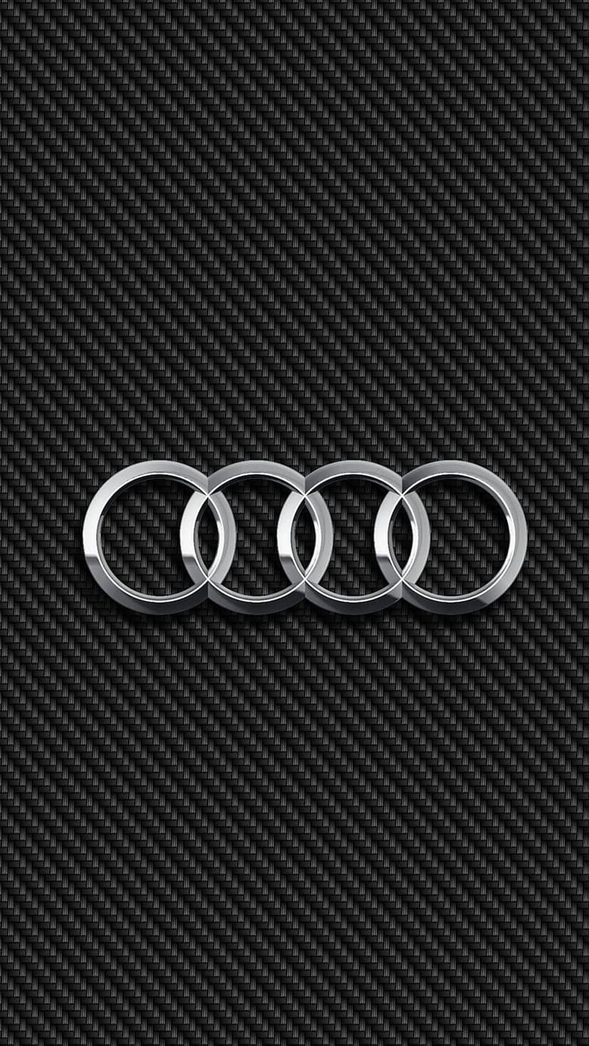 Silver Audi rings emblem centered on a textured black carbon fiber surface with sharp highlights and shadows.