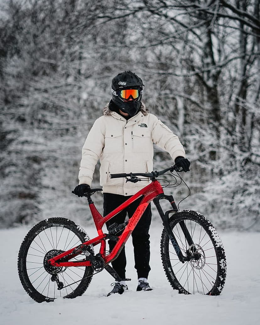 Mountain biker in a white jacket holds a red bike on a snowy trail surrounded by frost-covered evergreen trees.