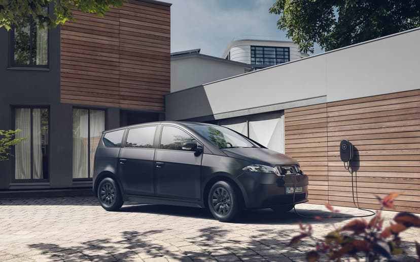 Matte black electric car charging at a modern house featuring wood paneling and a light cobblestone driveway.