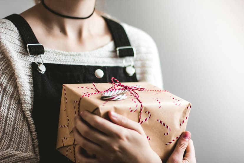 Hands holding a brown paper gift with red candy cane stripes, featuring a white knit sweater and black overalls.