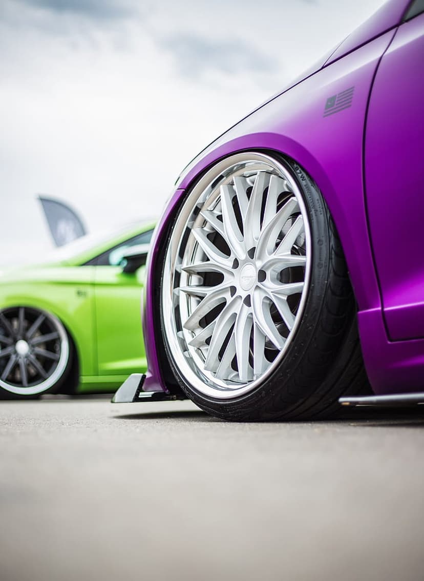 Silver alloy lowrider rims on purple and green customized cars show detailed chrome spokes and a lowered stance.