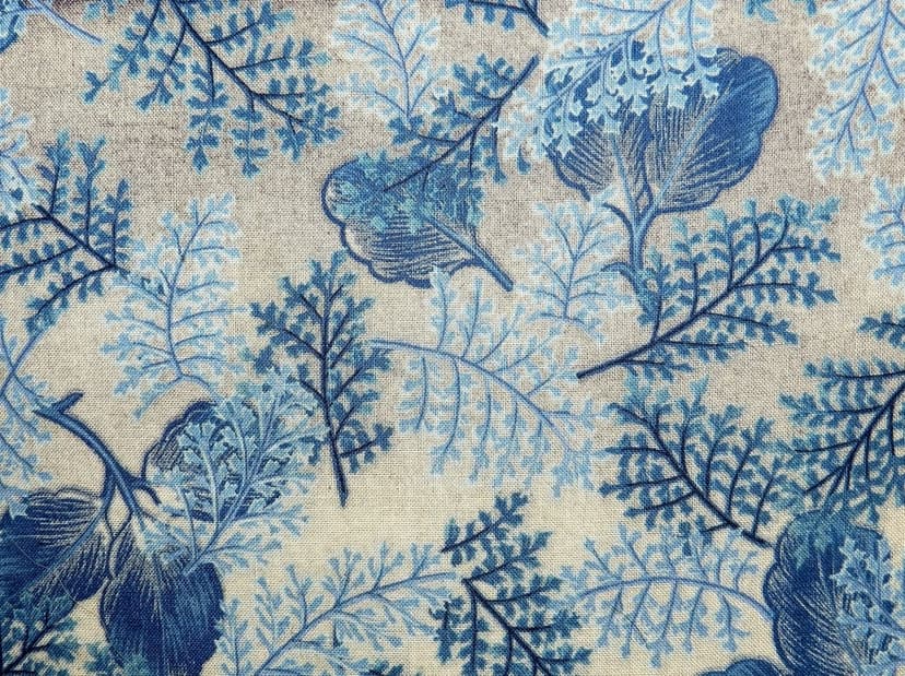 Blue fern fronds and stylized leaves overlap on a textured beige fabric surface with a natural fiber grain.